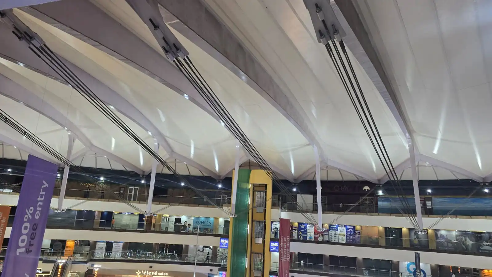 Vasant Square Mall Vasant Kunj — the underside of the translucent membrane lit from within the court at night, radial rib lines visible, Ekra Decor